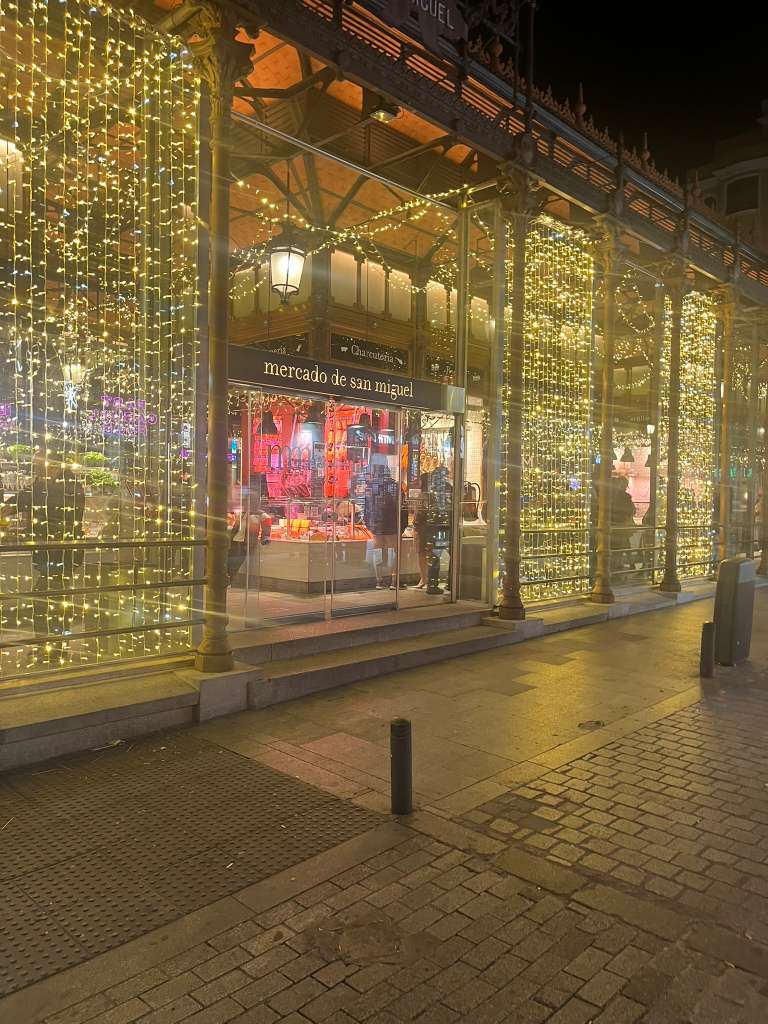 Glass market hall decorated with warm cascading string lights along a cobblestone street at night.