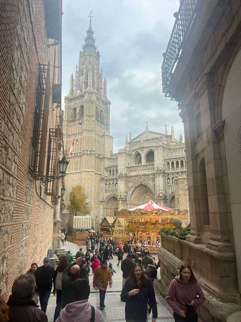 People strolling down a narrow stone street toward a cathedral tower and festive carousel.