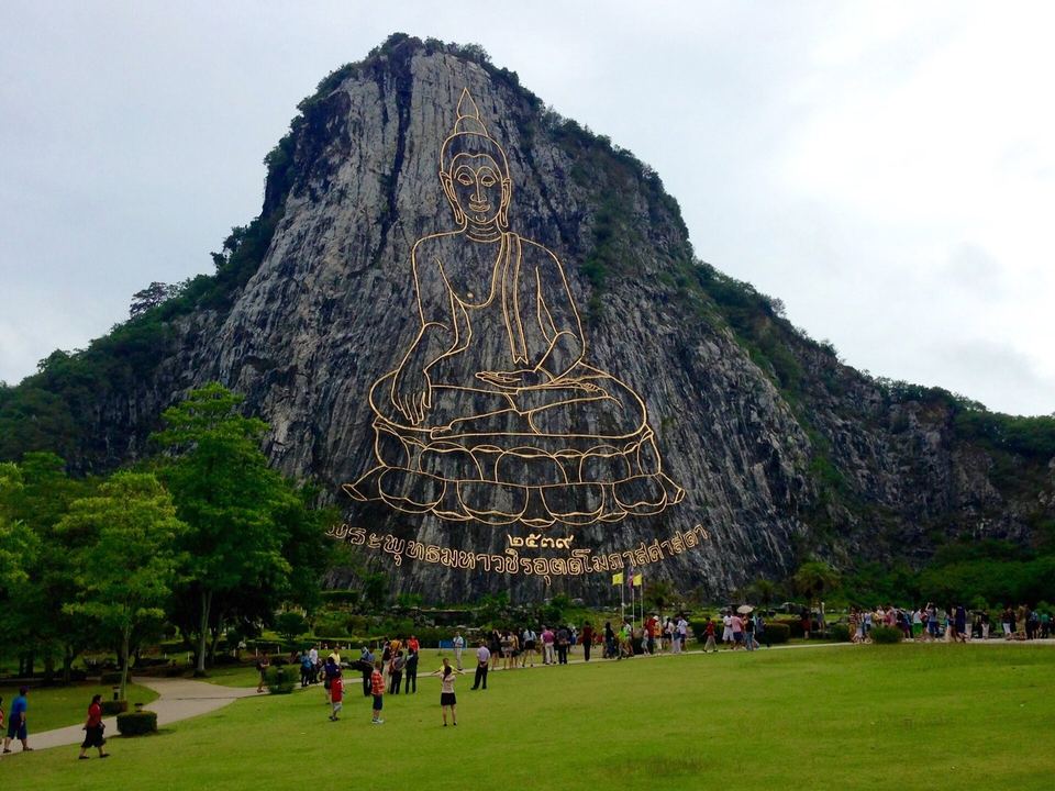 The gold Buddha figure of Khao Chi Chan etched into a limestone cliff above a lawn with visitors