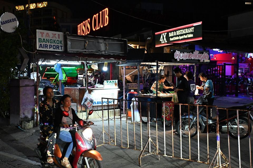 Roadside food stalls and neon bar signs on a Pattaya street at night, with motorbikes passing