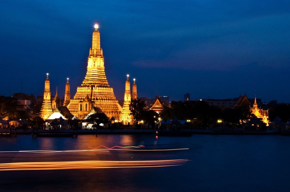 The floodlit spires of Wat Arun seen across the Chao Phraya River at night, with a boat trail on the water