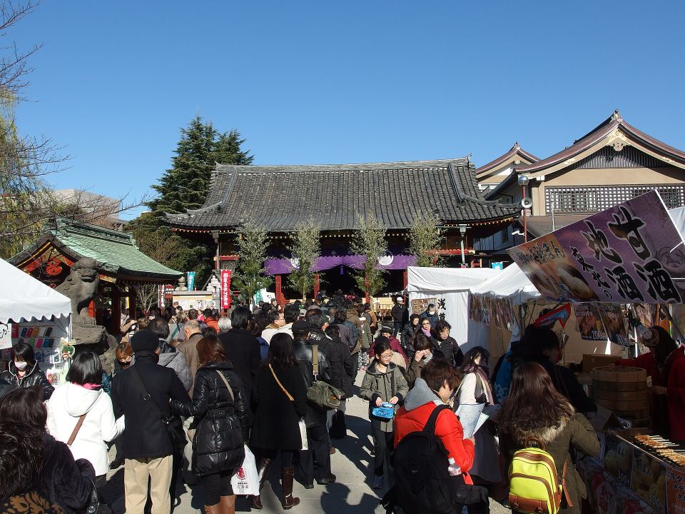 Crowds of people walk past outdoor food vendor stalls outside a Japanese temple.