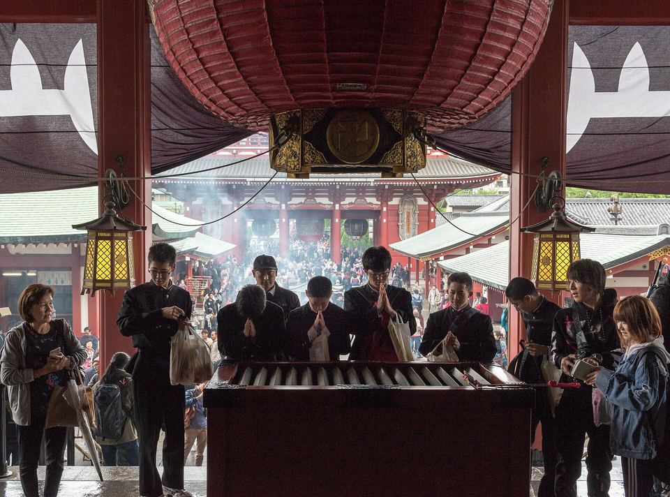 People pray at a wooden offertory box under a large red lantern at Senso-ji Temple.