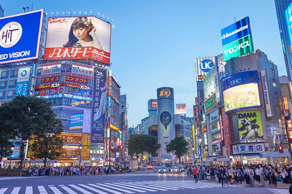Pedestrians wait at Shibuya Crossing surrounded by large digital billboards and commercial buildings.