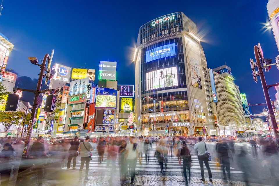 Blur of crowds walking across Shibuya Crossing in front of lit-up skyscrapers at night.