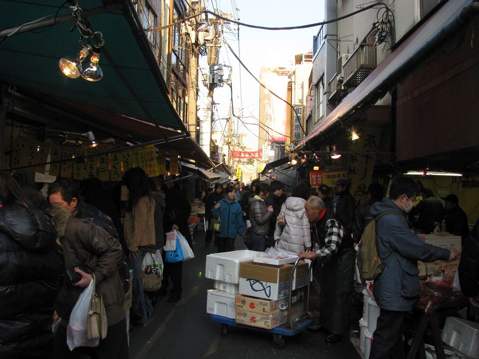 A vendor pushes a cart loaded with boxes down a narrow market alley crowded with shoppers.