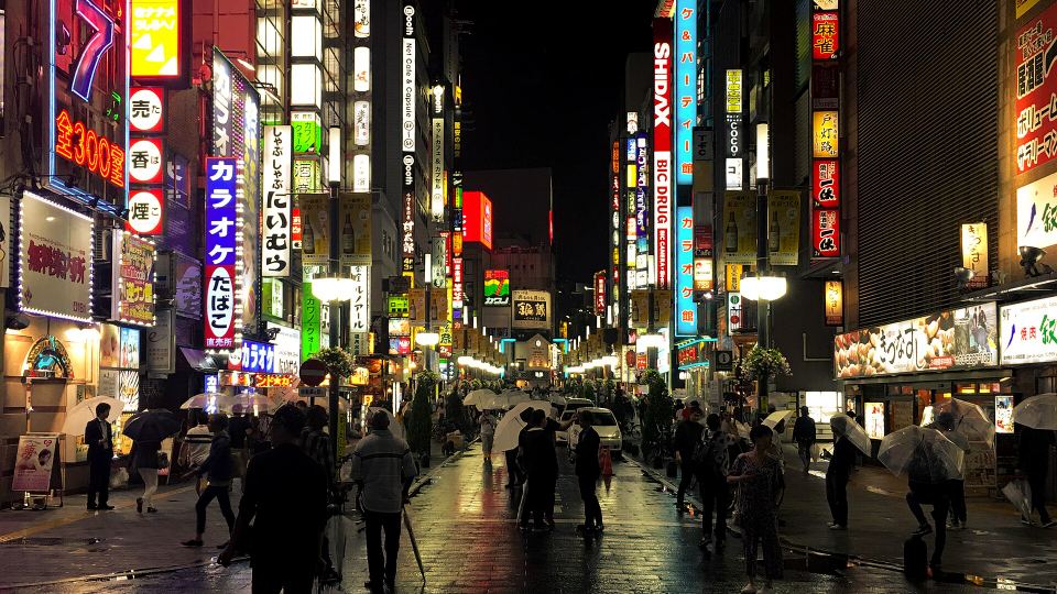 People carrying umbrellas walk down a rain-slicked alley lined with glowing Japanese neon signs.