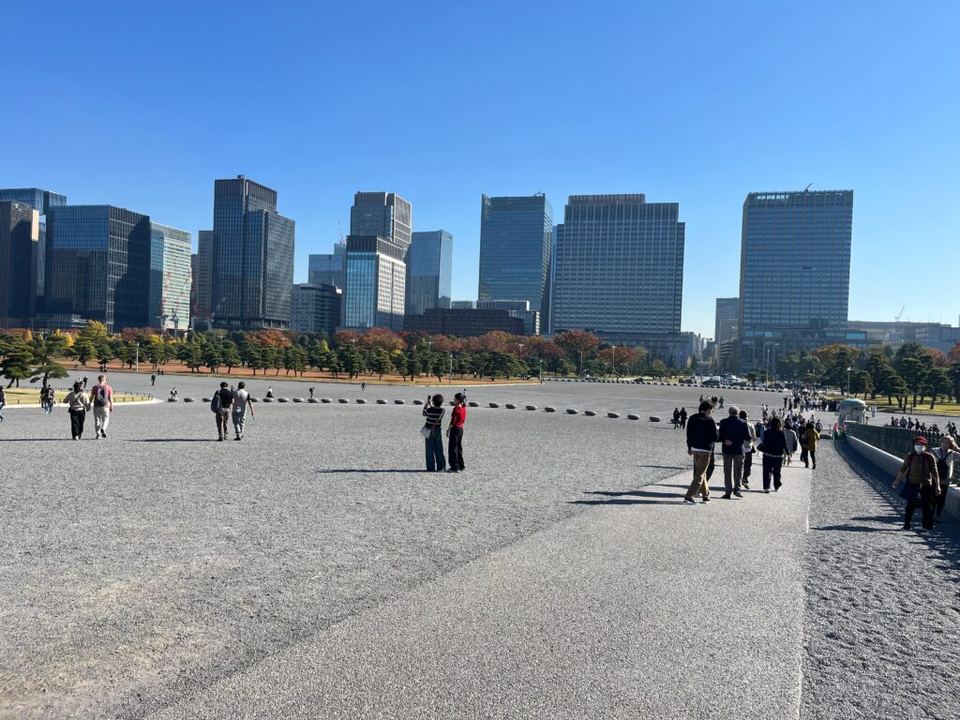 Pedestrians walking on a wide gravel plaza with tall modern office buildings in the background.