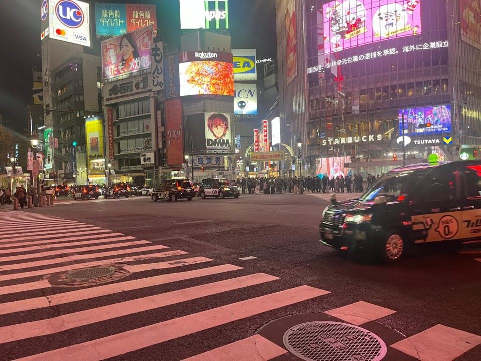 Night view of Shibuya Crossing with bright illuminated signs, crosswalk stripes, and moving taxis.