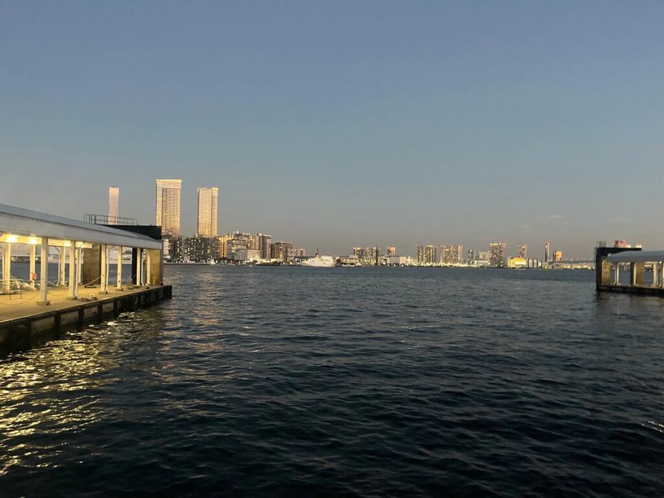 Calm water view from a pier toward an illuminated city skyline at twilight under a clear sky.