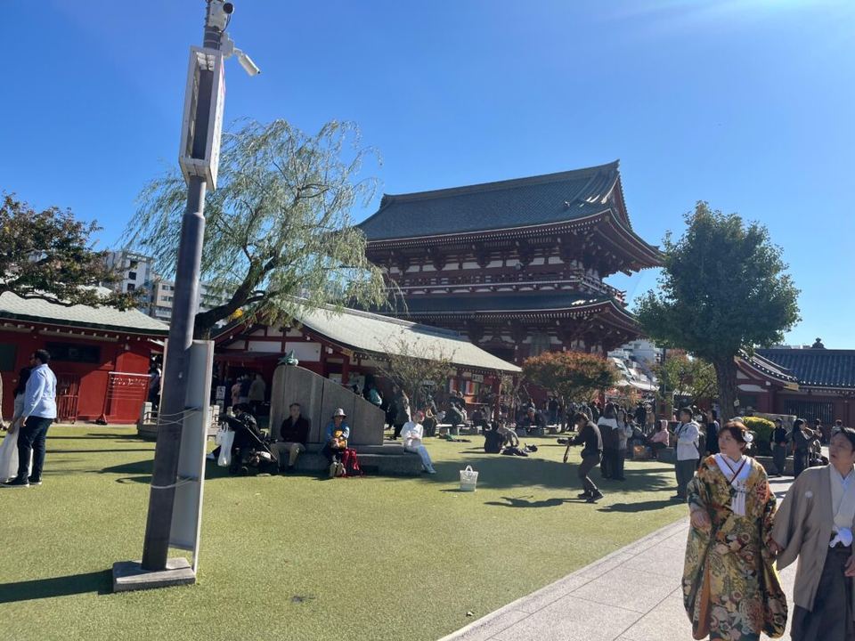 People walking past historic wooden temple buildings on a bright sunny day in Asakusa.